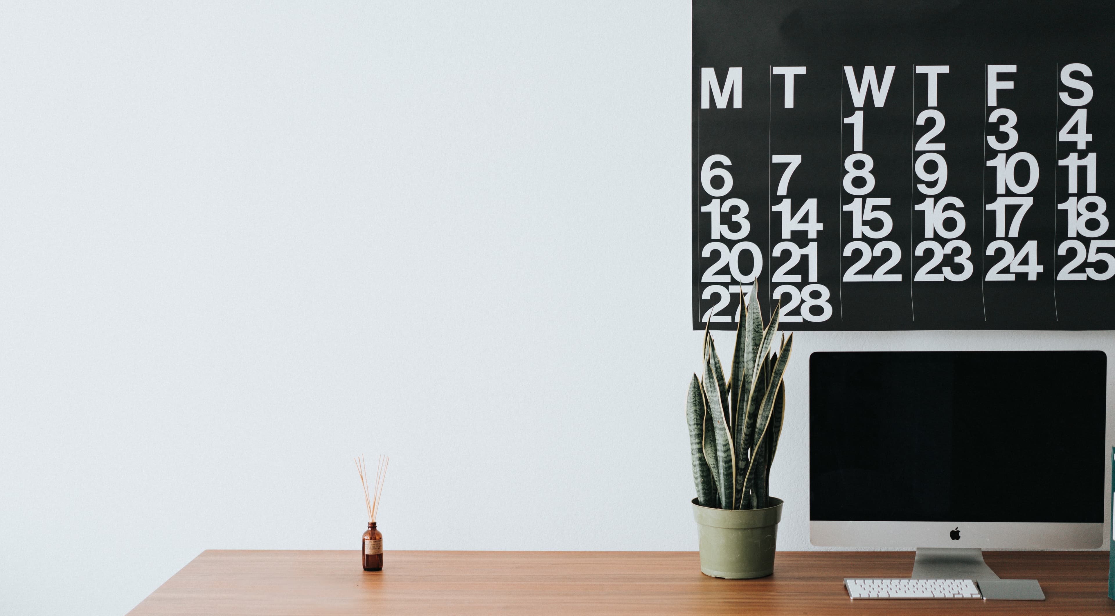 Minimalistic work desk with a large calendar hanging over a short plant and a computer on the right side of the desk
