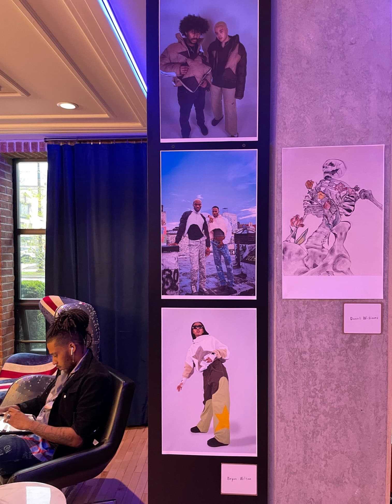 Man sitting in chair behind a wall with three photographs and one drawing hung up.